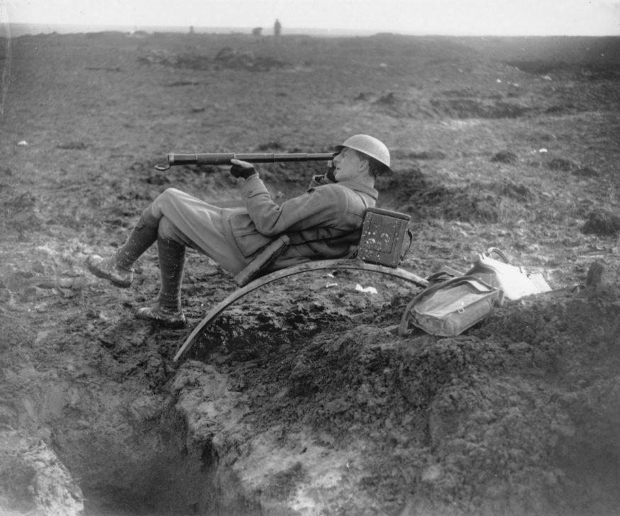 Australia's Official War Correspondent Charles Bean reclining in a muddy landscape, peering through a telescope