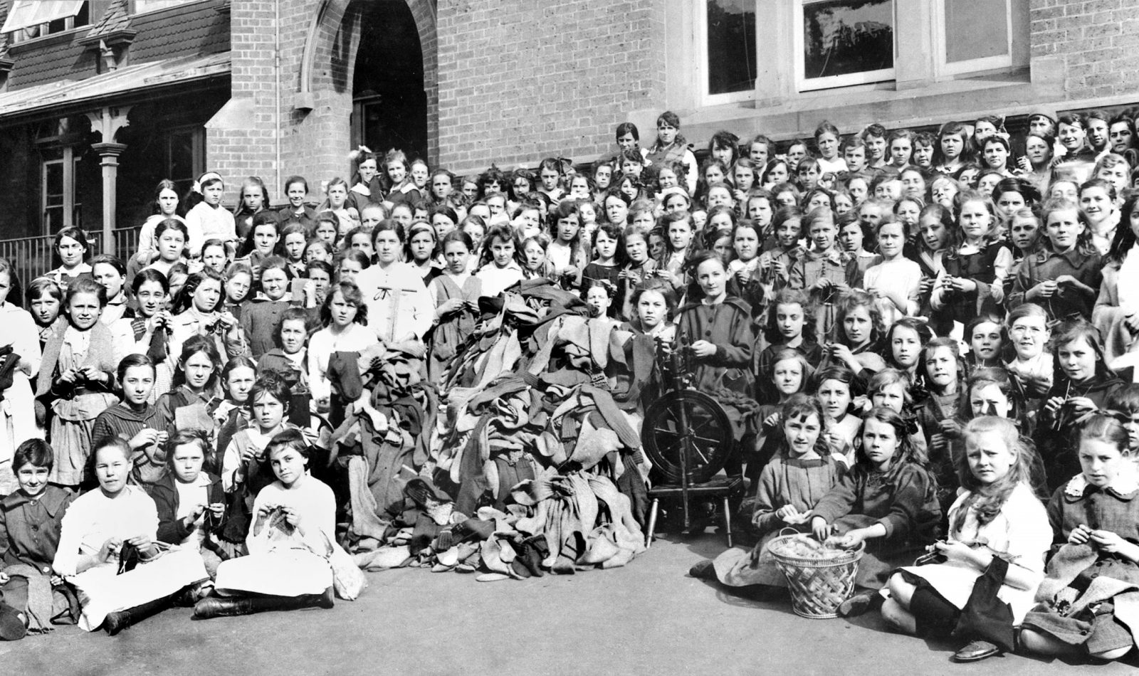 A large group of school children in front of red-brick building, assembled around a spinning wheel and a large pile of socks