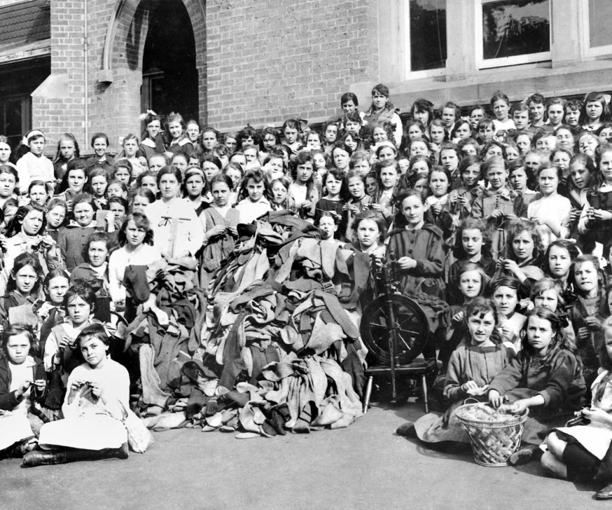 A large group of school children in front of red-brick building, assembled around a spinning wheel and a large pile of socks