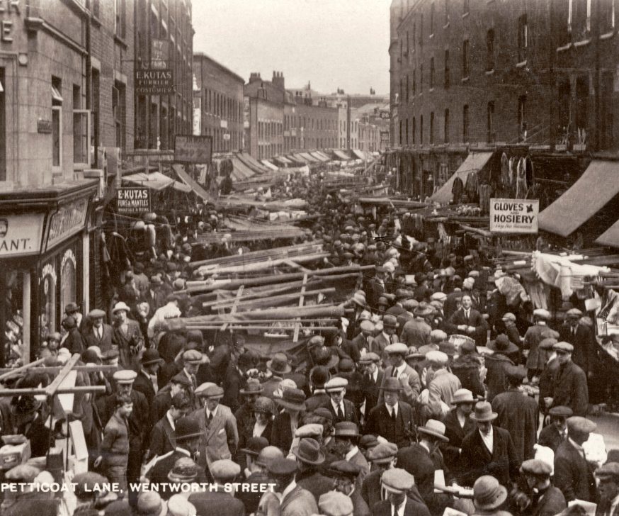 Petticoat Lane market, Wentworth Street, London early 1900s.  Jewish quarter, new immigrants from Eastern Europe.. Men all wearing caps and hats. Hebrew writing - Kosher restaurant. East End.